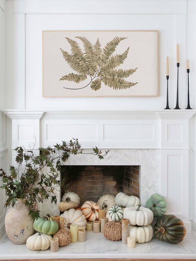 A fireplace with a large assortment of pumpkins covering the hearth in neutral fall colors. A piece of fall wall art hanging above.