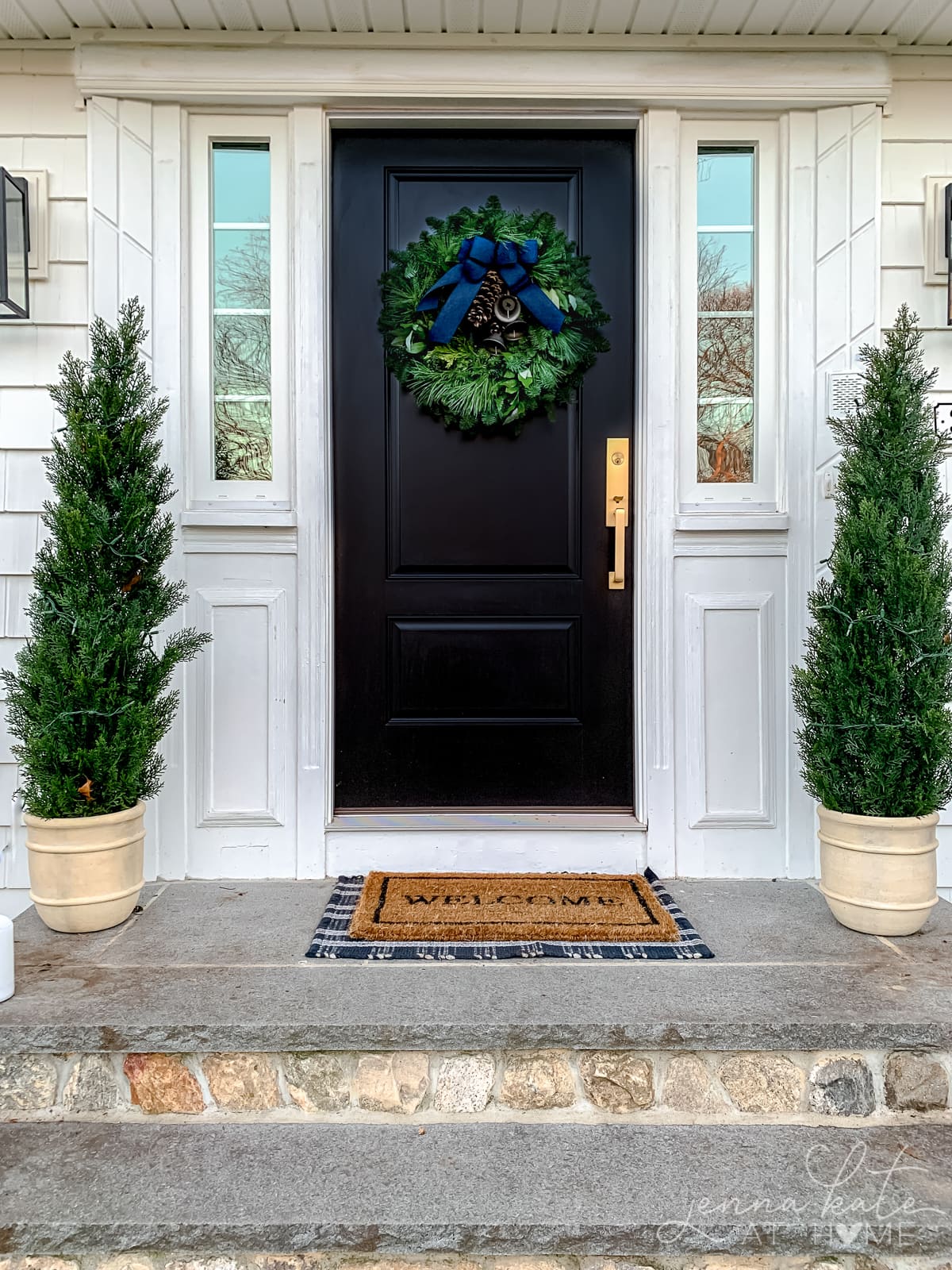 black front door with evergreen wreath with blue bow and cedar trees on either side