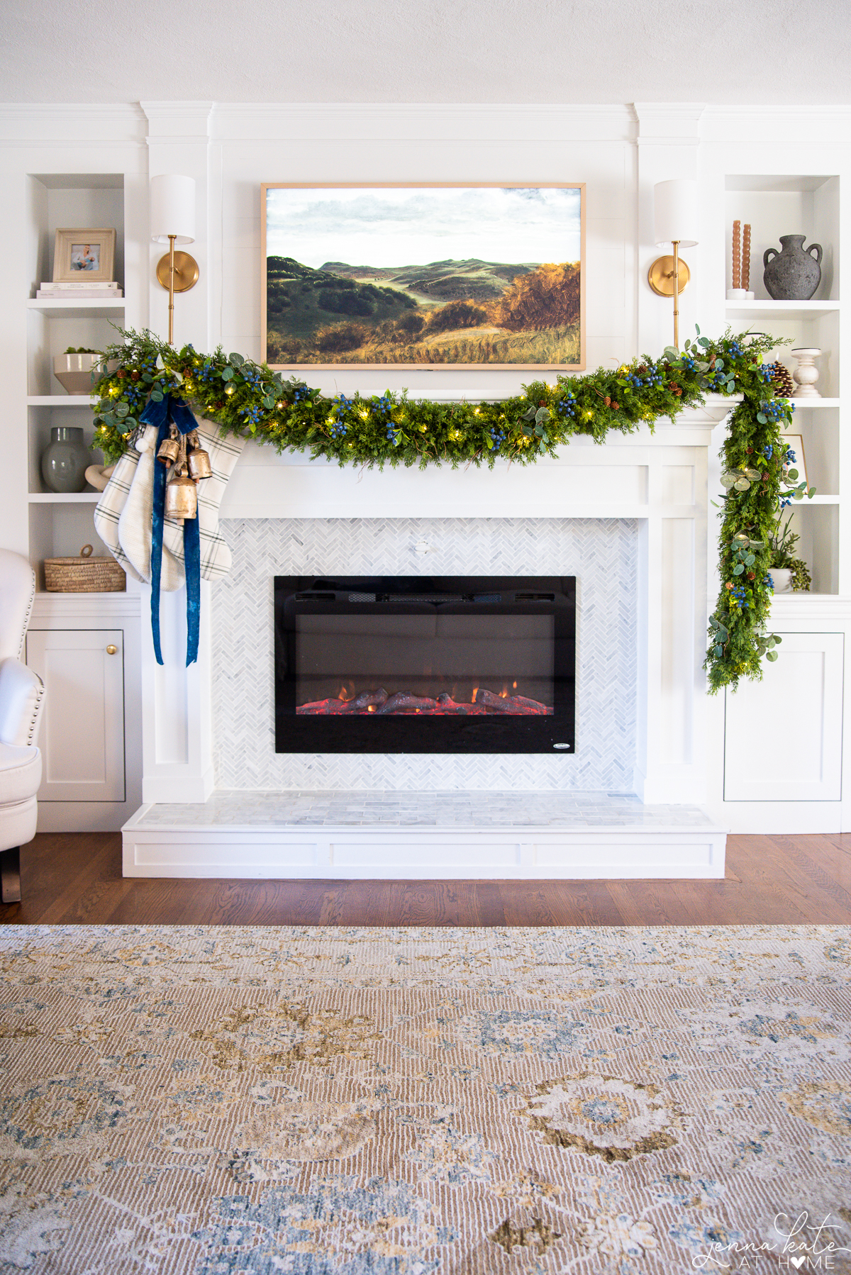 A view of a white fireplace and mantel decorated for Christmas with garland draped and gold bells hanging. Three stockings hang off the side.