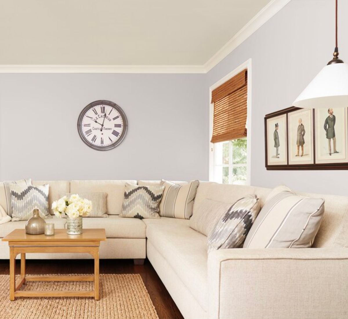 A living room with a sectional couch and walls painted with Sherwin Williams Grayish. A clock hung on the wall and wall art.