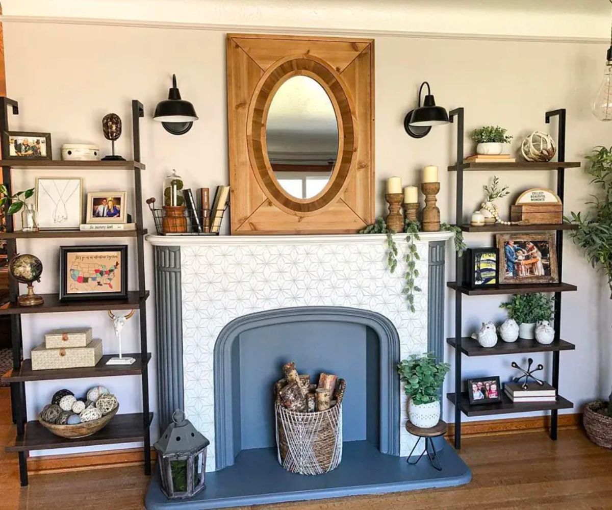 A fireplace wall decorated in a living room with wall color Grayish by Sherwin Williams. The fireplace is surrounded by two ladder bookcases decorated with stacked boxes, framed art, and a large mirror hanging overhead.