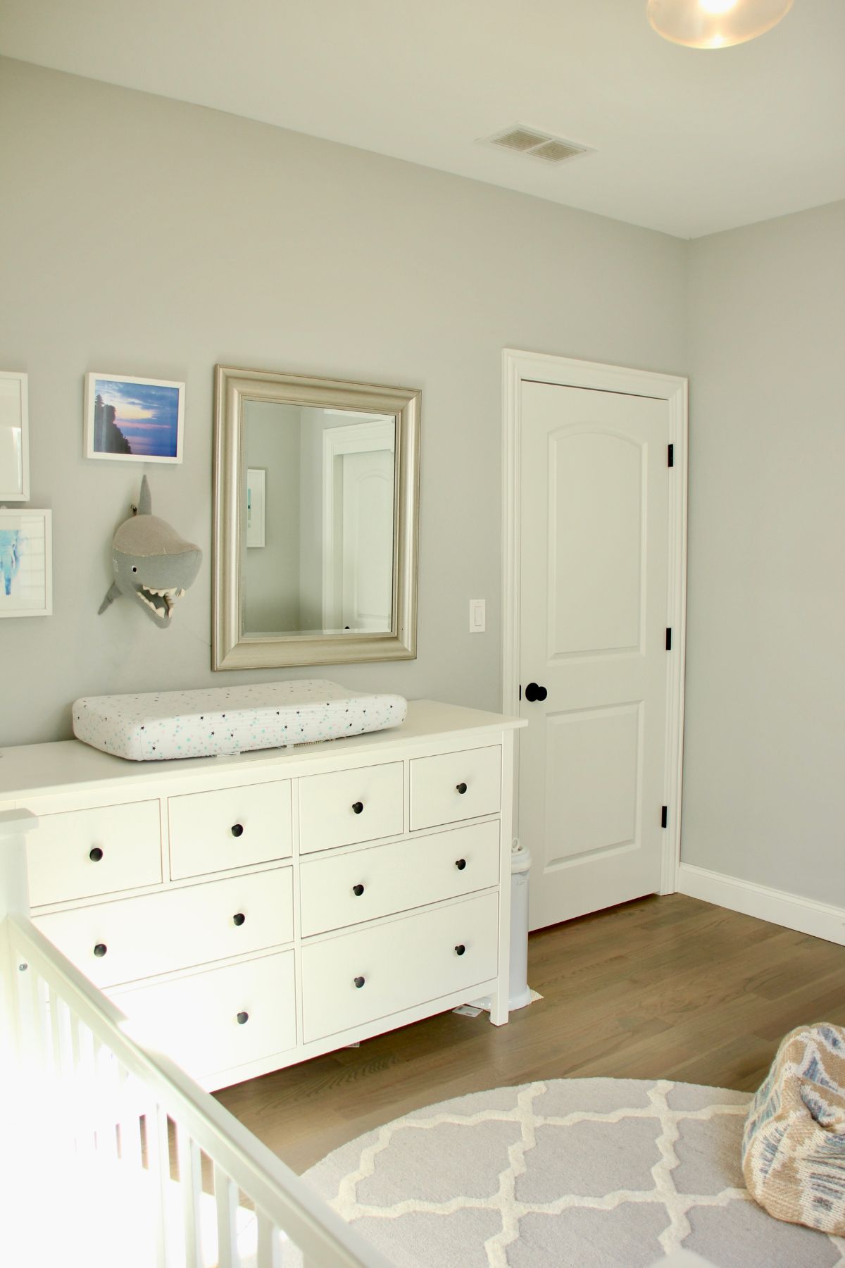 A neutral little boy nursery with a dresser beside the door and a mirror hanging overhead. A shark stuffed animal has been hung on the wall.