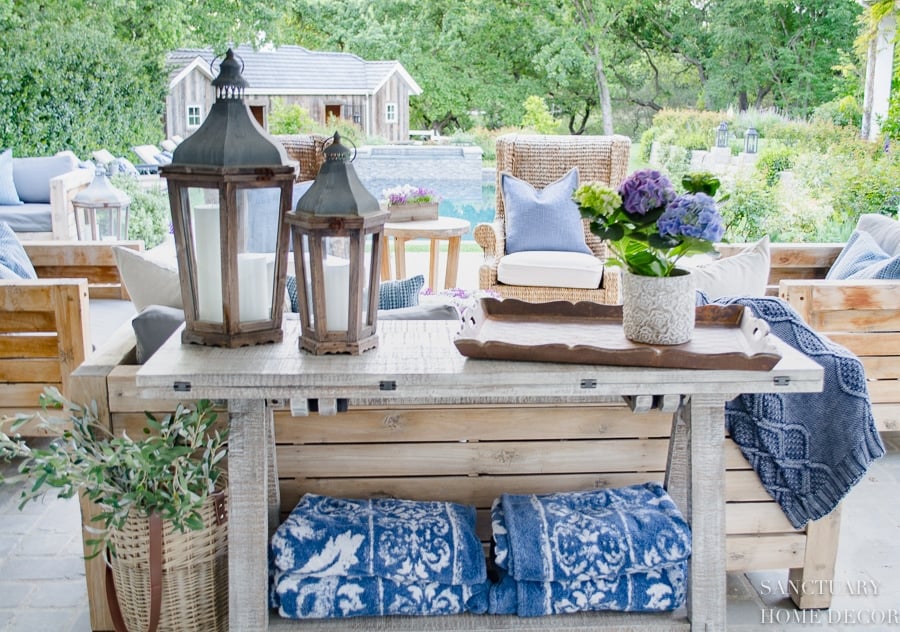 console table with a tray, potted plant and lanterns behind an outdoor couch.