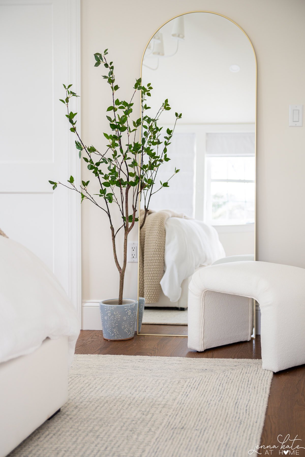 tall arched mirror with an ottoman and small artificial tree in front.