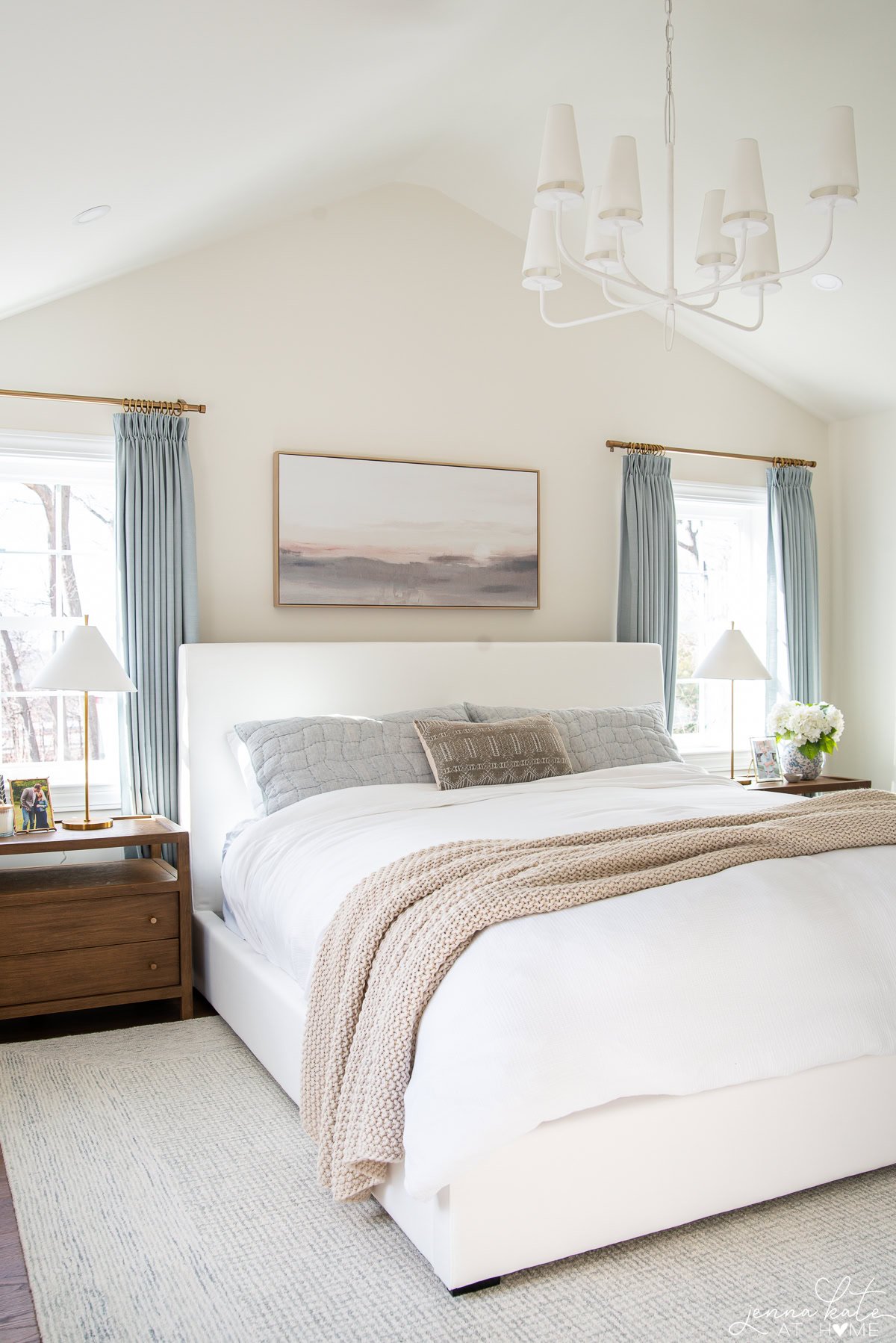 light snd airy bedroom with vaulted ceiling, white chandelier, white bed and blue curtains.