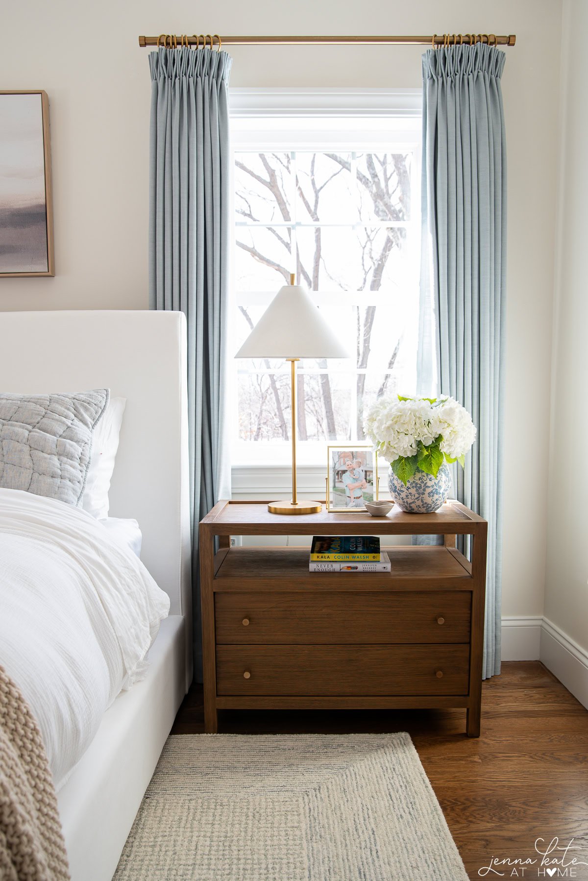 nightstand with table lamp and flowers and blue curtains flanking the window behind.