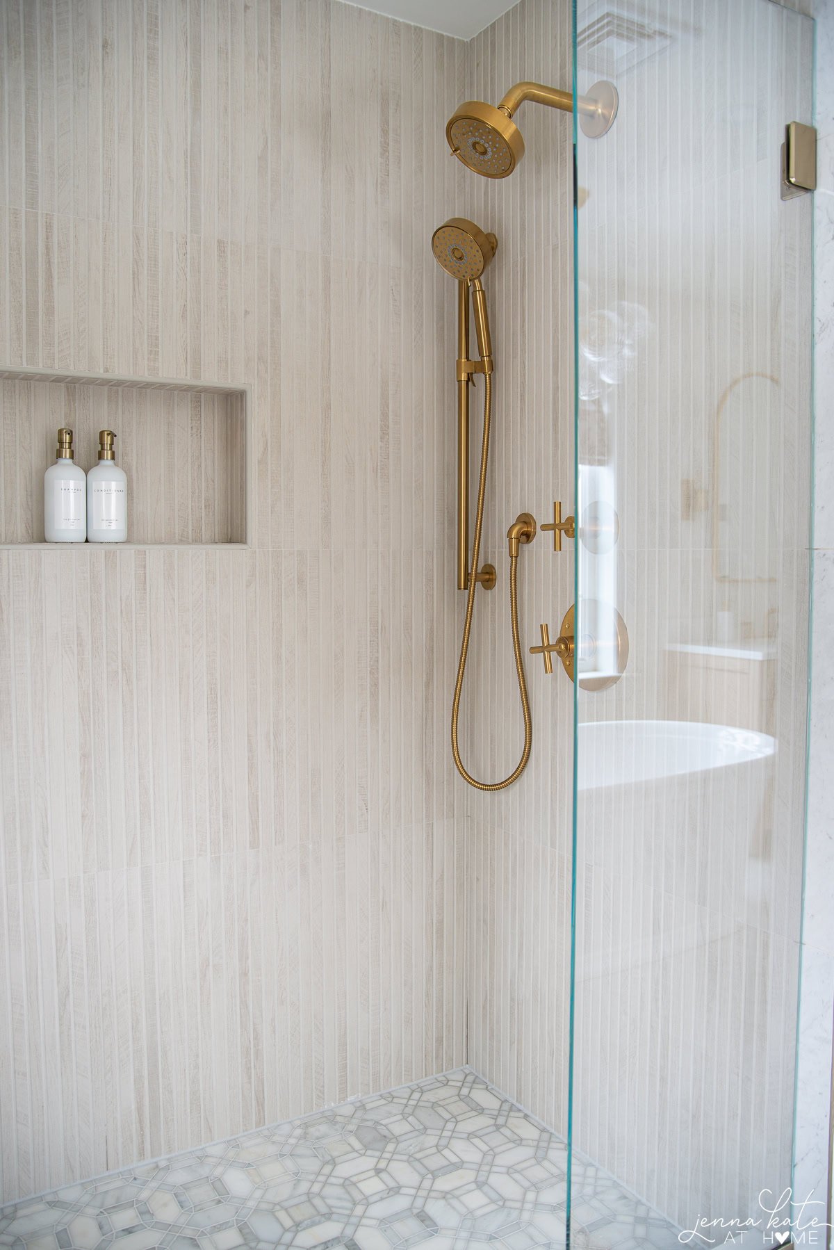 Walk-in shower with textured tile and brass Kohler fixtures in neutral bathroom.